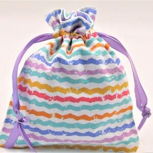May include: A drawstring bag with a colorful, wavy stripe pattern in shades of blue, purple, orange, yellow, and pink. The bag is tied with a lavender ribbon. The bag is made of fabric and is likely used for storage or gifting.