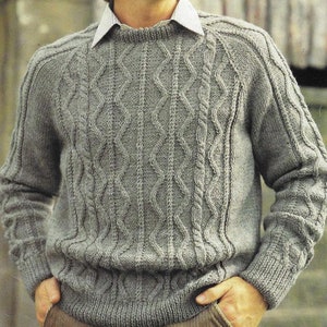 May include: A gray knitted sweater with a crew neck and long sleeves. The sweater features a cable knit design on the front and sleeves. The model is wearing a collared shirt underneath. The sweater is a medium weight and appears to be made of wool.
