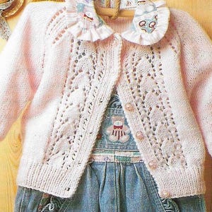 May include: A pink knitted cardigan with a lacy pattern and a frilled collar. The cardigan has a button fastening and is shown on a child's mannequin.