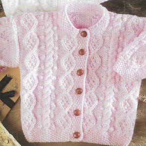 Könnte beinhalten: Ein rosa gestrickter Baby-Cardigan mit Zopfmuster und Knöpfen.
