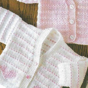 Op de afbeelding: Twee gebreide baby-vestjes, een wit met roze accenten en een roze met witte accenten. Beide vestjes hebben een knoopsluiting.