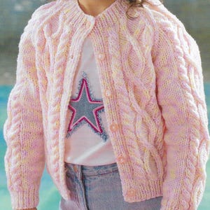 Peut inclure: Un cardigan en tricot rose et jaune à câble avec des boutons. Le cardigan est porté sur un t-shirt blanc avec un motif d'étoile bleue.