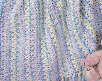 May include: A pastel-coloured crochet blanket with a fringe. The blanket features a chevron pattern in shades of blue, yellow, and pink. The soft, textured blanket is suitable for a baby or child.