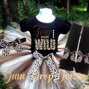 May include: A brown and white tulle tutu with a black shirt that says "Four ever Wild" in gold glitter. The tutu has a leopard print ribbon and bow. The shirt has a leopard print design on the bottom. The outfit is completed with black leg warmers with a leopard print bow.