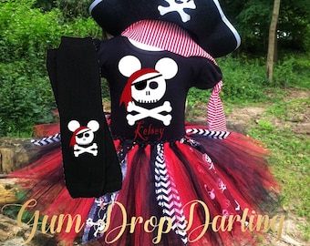 May include: A black pirate-themed outfit with a red and black tutu skirt, a black shirt with a skull and crossbones design, and a matching pirate hat. The outfit includes black leg warmers with a skull and crossbones design. The text "Gum Drop Darling" is visible.