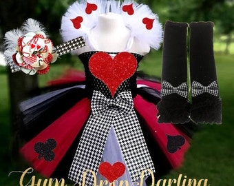 May include: A red, black, and white tutu with a red heart on the front. The tutu has a black and white houndstooth bow and a white tulle collar with red hearts. The set also includes a red and black card-themed headband and black leg warmers with a black and white houndstooth bow.
