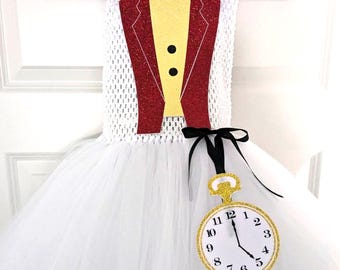 May include: White tutu dress with a red glitter jacket, yellow vest, and red bow tie. The dress features a gold glitter pocket watch with black hands. The costume is hanging on a white hanger.