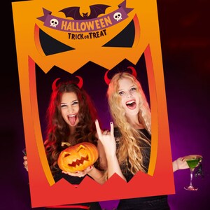 Könnte beinhalten: Zwei Personen, die als Teufel verkleidet sind, posieren für ein Foto durch einen Halloween-Fotobooth-Ausschnitt. Der Ausschnitt ist orange und rot mit einem Kürbisgesicht und den Worten "Halloween Trick or Treat". Eine Person hält einen geschnitzten Kürbis und die andere ein grünes Getränk.