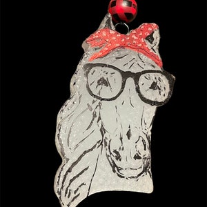 May include: White horse head ornament with black and white details, wearing red and white bandana and black framed glasses. The ornament is hanging from a black string with a red and black checkered bead.