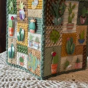 May include: A square box constructed from panels of yarn-stitched plastic canvas in shades of green, yellow, and cream. The exterior is decorated with numerous small, flat, colorful stickers depicting various cacti and succulents in pots.