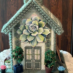 May include: A decorative house crafted from sage green plastic canvas with stitched details. A large paper succulent is centered on the front facade above a stitched door. Miniature potted plants with red and orange berries flank the entrance.