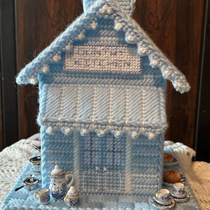 May include: A light blue plastic canvas house with a chimney and a sign reading COUNTRY KITCHEN. The structure features a roof with white bead accents, a front awning, and a base decorated with miniature ceramic kitchenware and food items.