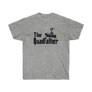 May include: A heather gray t-shirt with the text "The Quadfather" in black, accompanied by a graphic of four drums. The design is centered on the front of the shirt. The shirt has a crew neck and short sleeves.