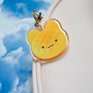 May include: A yellow and white cartoon character keychain shaped like a pastry with a smiling face. The keychain is made of clear acrylic and has a gold metal ring.