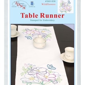 May include: A white table runner with a stamped embroidery design of pink and purple wildflowers with butterflies. The table runner is 15 inches by 42 inches and is made of cotton and polyester broadcloth. The text "#560-838 Wildflowers Table Runner Stamped for Embroidery" is printed on the packaging.