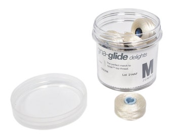 Może przedstawiać: Przezroczysty plastikowy pojemnik z białą pokrywką wypełniony szpulami kremowej nici. Pojemnik zawiera tekst "gna-glide delights" i "The perfect match to Glide 100 thread."  Pojemnik zawiera również tekst "CREAM" i "Lot 214AF".  Pojemnik ma etykietę z literą "M" i tekstem "JUMBO".