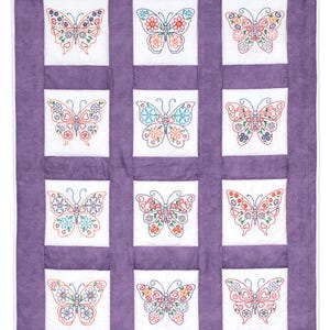 Puede incluir: Una colcha morada con nueve cuadrados blancos, cada uno con una mariposa bordada de colores con diseños florales y de corazón.