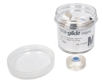 Może przedstawiać: Przezroczysty plastikowy pojemnik z białą pokrywką wypełniony białymi szpulami nici. Pojemnik zawiera tekst "magna-glide delights" i "The perfect match to Glide top thread." Pojemnik zawiera również tekst "Lot 11121AF" i "M JUMBO".