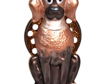 Puede incluir: Una figura de perro caprichosa con una combinación de colores bronce y marrón oscuro. El perro tiene ojos grandes y expresivos y está sentado. Un elemento decorativo de color dorado está detrás del perro.
