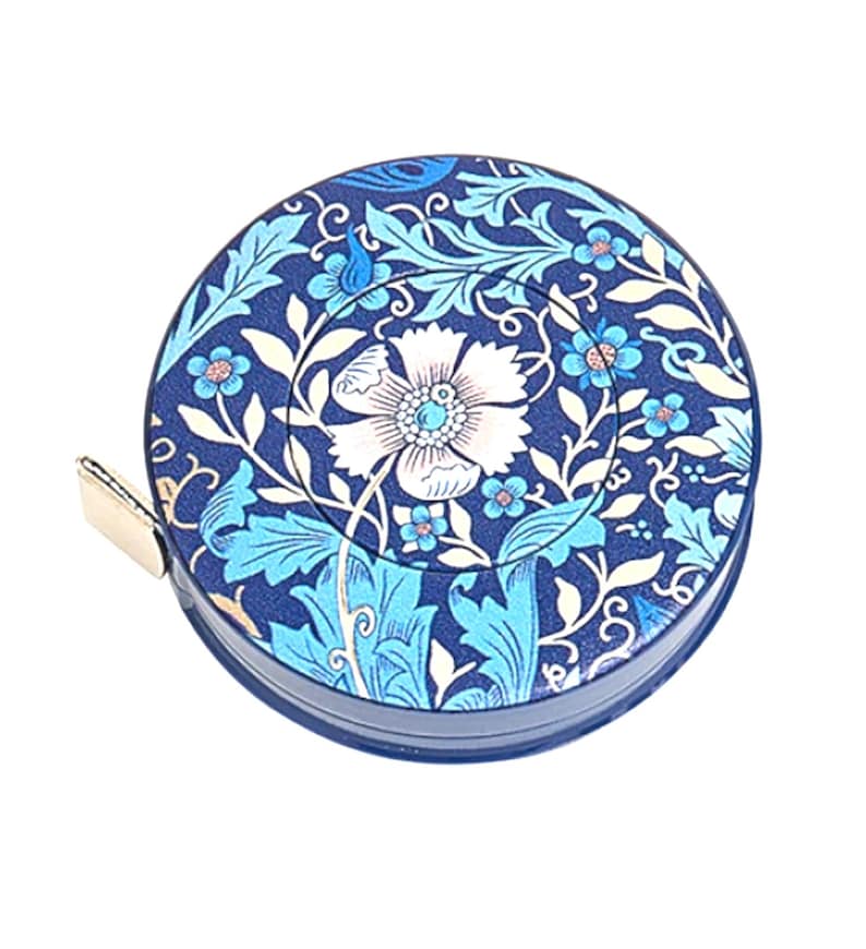 May include: A circular navy blue tape measure with a floral pattern. The design features a large white flower at the centre, with blue and light blue leaves and flowers. A gold-coloured metal tab is visible on the side.
