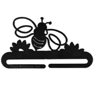 May include: Black metal bee-shaped decorative item. The bee is silhouetted with detailed wings and antennae, perched above stylized flowers. The bottom section has a slot and the text "Patch Abilities, Inc."