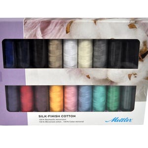 May include: A rectangular box containing sixteen spools of Mettler Silk-Finish Cotton thread in various colors, arranged in two rows. The packaging features a lavender side panel and a graphic of cotton bolls, with the text Silk-Finish Cotton.
