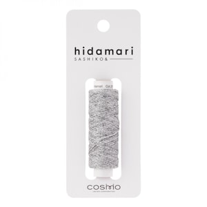 Op de afbeelding: Een spoel grijs garen met het label "hidamari SASHIKO &" en "Col.22". Het garen is verpakt in een doorzichtige plastic verpakking met een wit label waarop "cosimo LECIEN CORPORATION" staat.