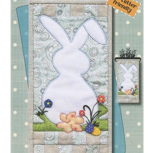 Könnte beinhalten: Ein Quiltmuster für eine Wandbehänge in Form eines Hasen. Das Muster ist 15,2 cm x 30,5 cm groß und für Schneidemaschinen geeignet. Das Muster zeigt eine weiße Hasensilhouette mit grünem Gras und Blumen.