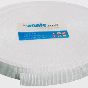 May include: A large, circular spool of white woven webbing tape. A rectangular label is centered on the top surface, featuring the text byannie.com, what's in your bag?, a phone number, email address, website, and various social media icons.