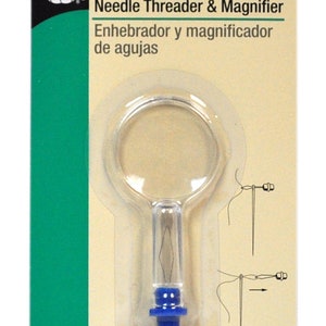 Puede incluir: Un enhebrador de agujas y una lupa de plástico transparente con un mango azul. El enhebrador está empaquetado en una tarjeta beige con el texto "Needle Threader & Magnifier" y "Enhebrador y magnificador de agujas".