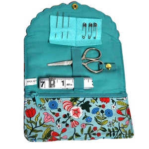 May include: Open teal sewing kit with a floral patterned exterior. The kit contains needles, safety pins, small scissors, and a 60-inch measuring tape. The kit is secured with a zipper and a snap closure.
