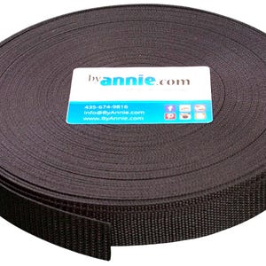 May include: A large, circular roll of dark brown woven webbing tape. A rectangular white and blue card is placed on top, featuring the text byannie.com, the phone number 435-674-9816, the email info@ByAnnie.com, and the website www.ByAnnie.com.