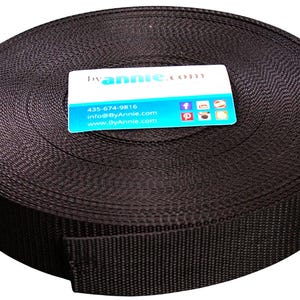 May include: A large, circular roll of dark brown woven nylon webbing tape. A rectangular white and blue business card is placed on top of the roll, featuring the text byannie.com, the phone number 435-674-9816, and various social media icons.