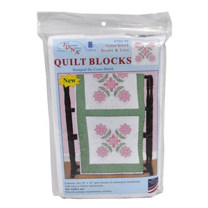 May include: A packaged cross-stitch kit labeled "Quilt Blocks" with the text "Hearts & Lace." The kit features a design of pink floral patterns on white fabric, with green borders. The package includes six 18" x 18" quilt blocks.