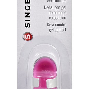 Pode incluir: Um dedal de gel rosa da marca Singer com uma banda de conforto branca. A embalagem diz "Comfort Fit Gel Thimble" e inclui texto em espanhol e francês. O número do produto é 07330.