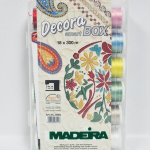 Może przedstawiać: Przezroczyste plastikowe pudełko zawierające 10 szpule nici do haftu Madeira Decora Smart Box. Szpule są w różnych kolorach, w tym czerwonym, różowym, niebieskim, żółtym, zielonym i białym. Pudełko jest oznaczone nazwą marki Madeira i nazwą produktu Decora Smart Box. Tekst na pudełku podaje również, że nić ma długość 18 x 300 m.