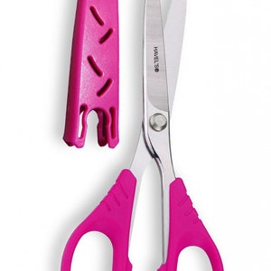 May include: Bright pink scissors with a matching protective cover. The scissors have a silver blade and pink handles. The brand name "HAVELS®" is printed on the blade. The cover has a series of cut-out shapes.