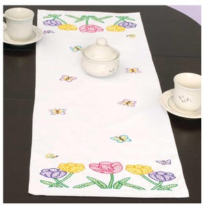 May include: White table runner with embroidered colorful tulips and butterflies. The runner is on a wooden table with two white cups and saucers.