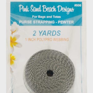 May include: A roll of pewter-colored polypro webbing for bags and totes, packaged in a clear plastic bag. The label reads Pink Sand Beach Designs #506, 2 Yards, 1 Inch Polypro Webbing, and Purse Strapping - Pewter, with white flower graphics.