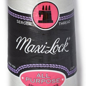 Puede incluir: Un carrete blanco de hilo para overlock Maxi-Lock. La etiqueta es negra con texto blanco y un círculo rosa con un icono de máquina de coser. El hilo es de poliéster hilado multiusos.