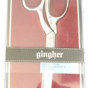 May include: Silver-colored dressmaker's shears in a clear plastic case with a red background. The shears have a knife edge and the brand name "gingher" is printed on the packaging.