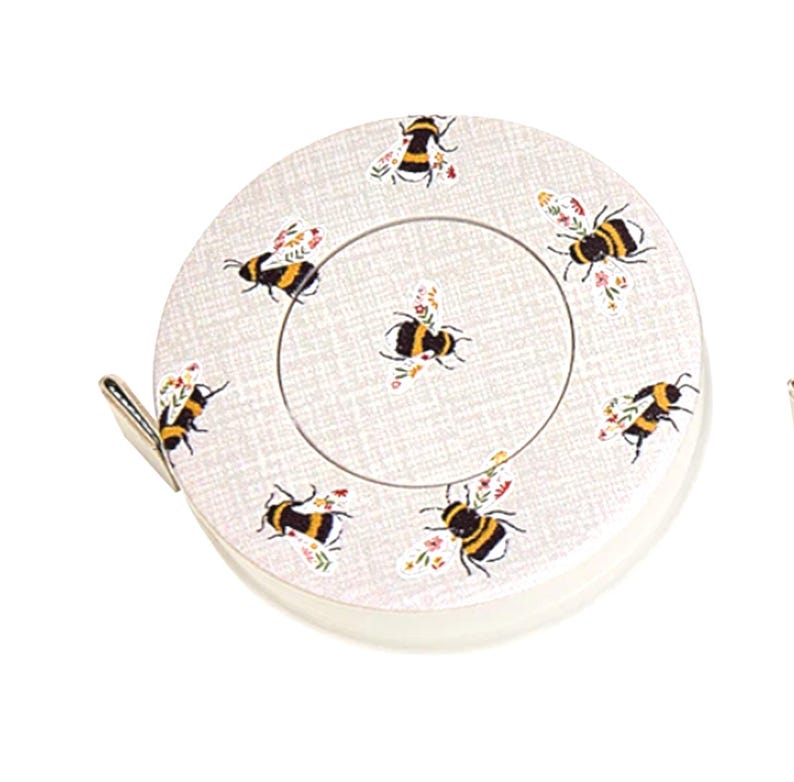 May include: A circular tape measure with a bee and floral design. The tape measure is off-white with a metal clip and a white border. The bees are black and yellow with floral details. A practical tool for measuring in sewing and craft projects.