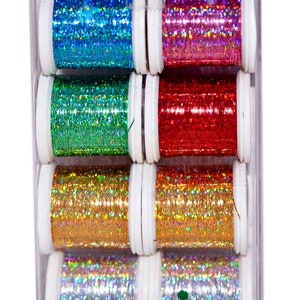 May include: A clear plastic case containing eight spools of glitter thread in various colours: blue, pink, green, red, gold, and silver. The spools are white with the brand name "MADEIRA" in green. Suitable for crafting and embroidery projects.
