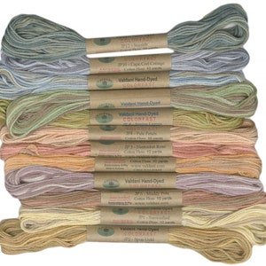Puede incluir: Una pila de 10 ovillos de hilo de algodón teñido a mano Valdani en varios tonos de verde, rosa, naranja y amarillo. Cada ovillo está etiquetado con el nombre y el número del color, y el logotipo de Valdani. El hilo es resistente a la decoloración y perfecto para el bordado y otras manualidades.