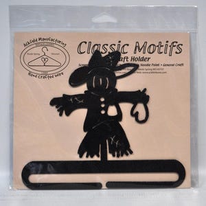 May include: A black metal scarecrow-shaped craft holder by Ackfeld Manufacturing, labeled Classic Motifs. The silhouette features a wide-brimmed hat, a jacket with buttons, and a horizontal bar base for hanging items, packaged in a clear plastic sleeve.