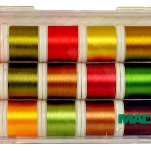 Puede incluir: Una caja de plástico transparente que contiene 15 carretes de hilo de bordar Madeira. Los carretes están dispuestos en tres filas de cinco. Los colores del hilo son naranja, amarillo, verde, marrón y rojo.