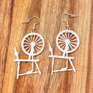 May include: A pair of white, laser-cut earrings. Each earring features a spinning wheel design with a circular wheel and a rectangular base. The earrings hang from silver-colored fish hook ear wires. The background is a wooden surface.