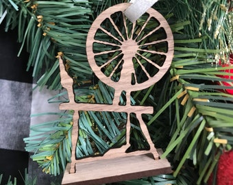 May include: Wooden Christmas ornament shaped like a spinning wheel. The ornament is brown and has a white ribbon attached to it.
