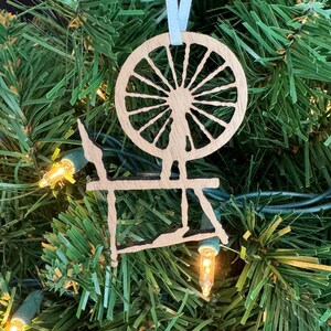 May include: A wooden spinning wheel ornament with a light blue ribbon hanging from the top. The ornament is a silhouette of a spinning wheel with a large wheel and a smaller base.