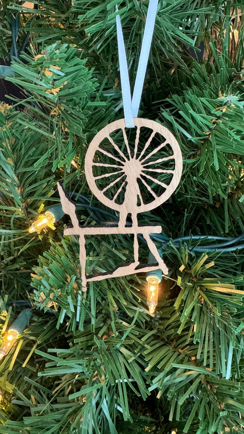 May include: A wooden spinning wheel ornament with a light blue ribbon hanging from the top. The ornament is a silhouette of a spinning wheel with a large wheel and a smaller base.
